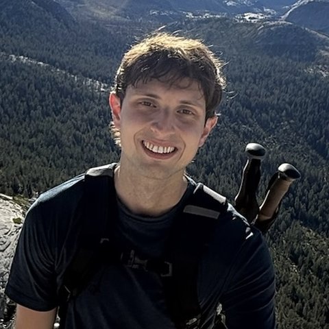 Matthew smiling on a hike, with mountains and forest behind him